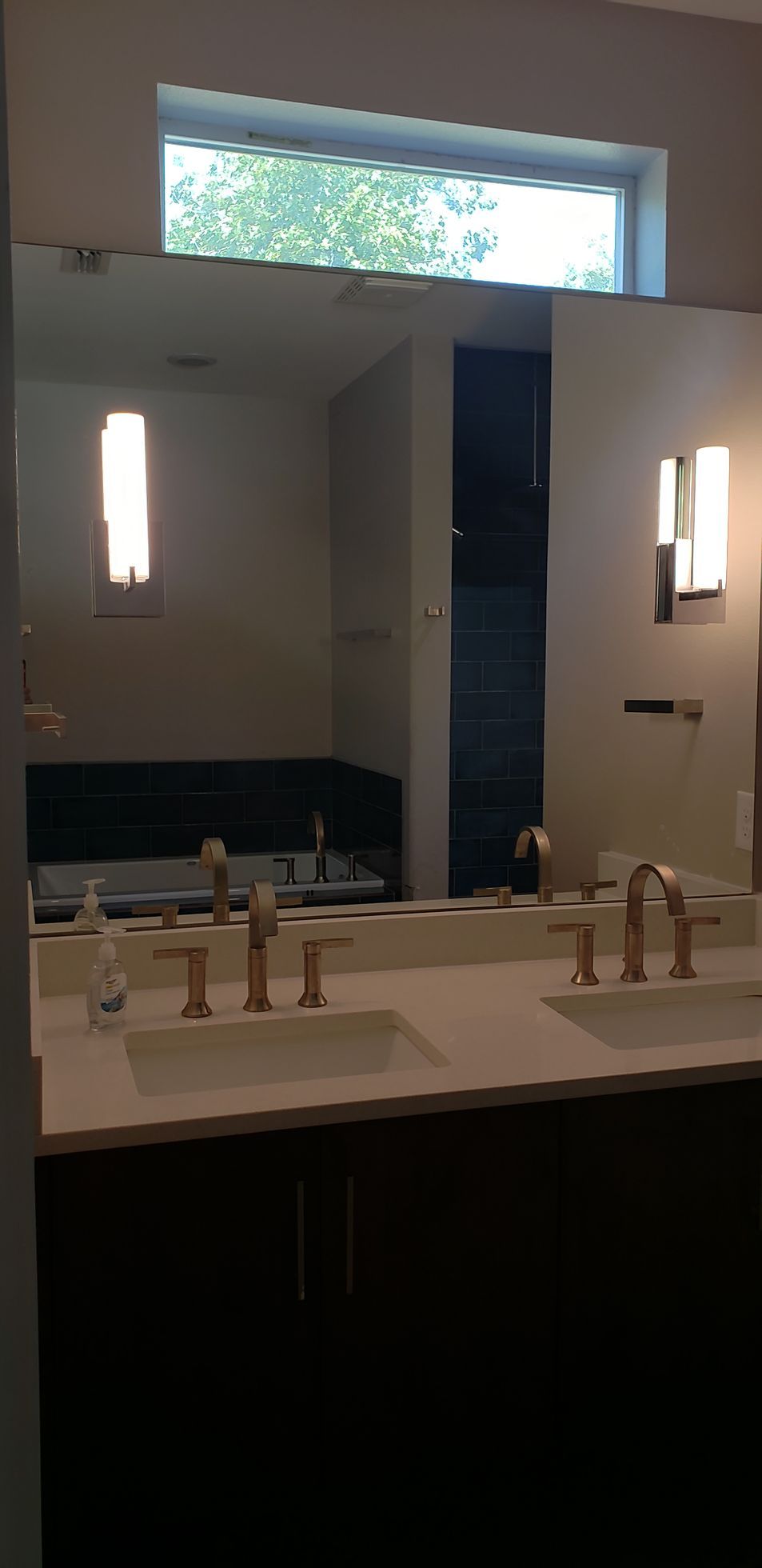 Bathroom with double sinks, large mirror, and wall sconces. Dark cabinets, white countertop. Natural light from a window.