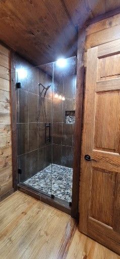 Rustic-style shower with glass door and pebbled floor, in a wood-paneled bathroom.