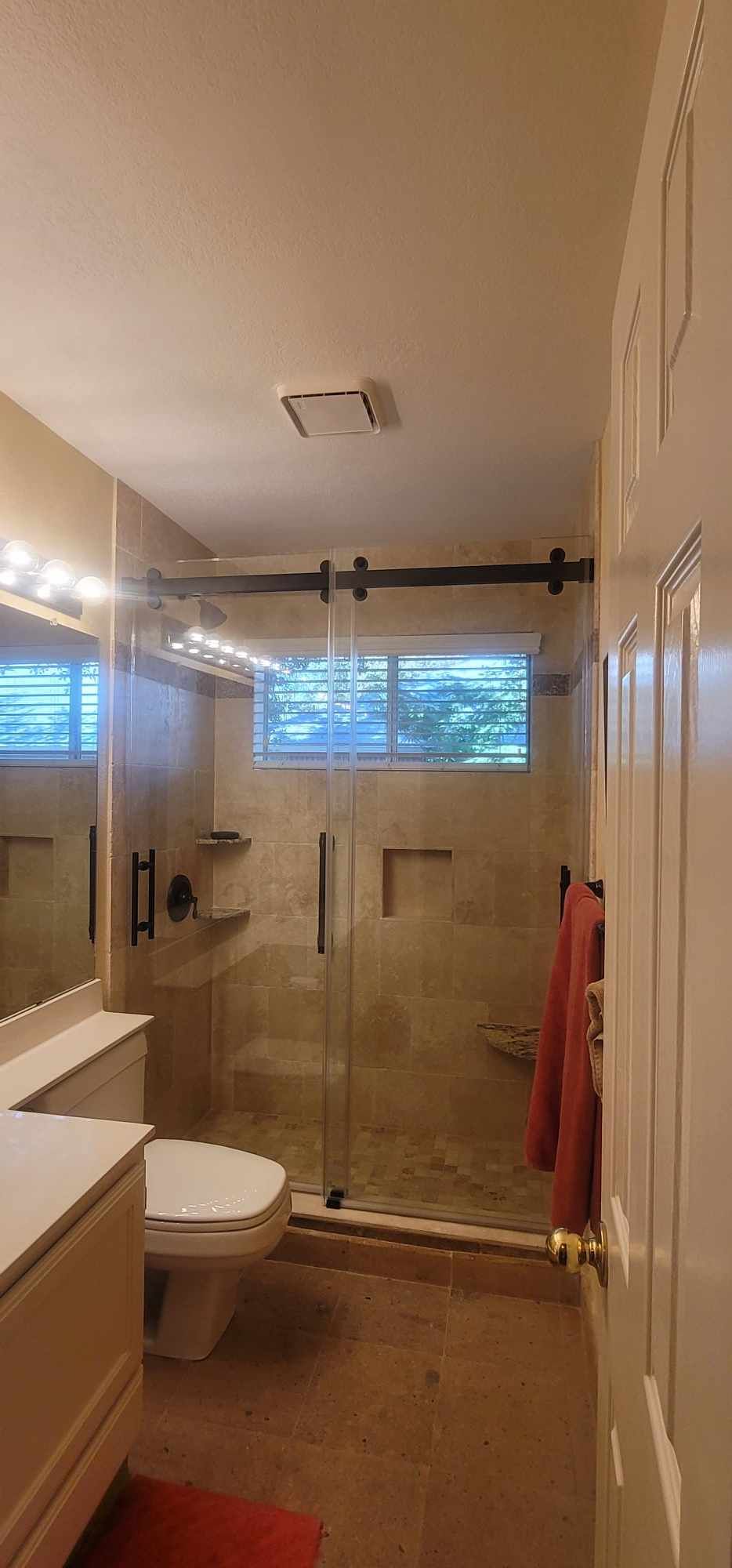 Bathroom with glass shower, toilet, and vanity. Beige tile walls and red bath mat.