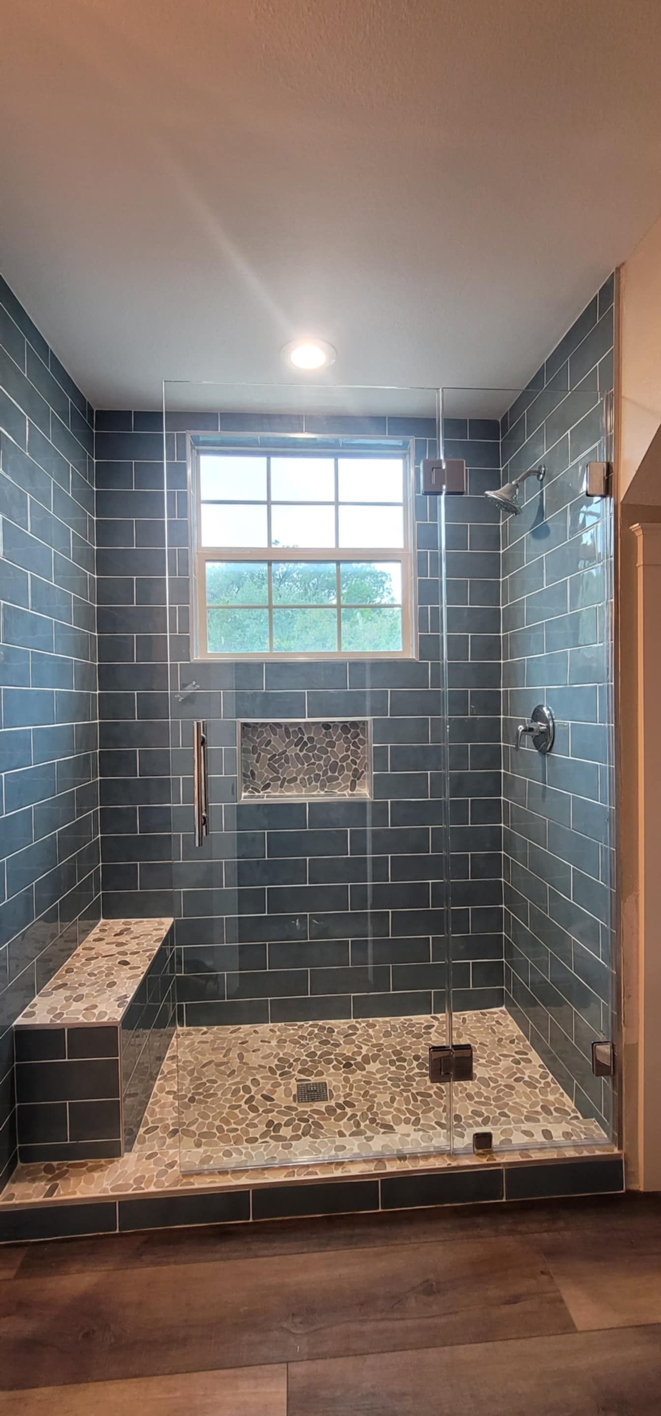 Modern shower with dark blue-gray tiled walls, a pebble floor, glass doors, and a built-in bench. A window is in the back.