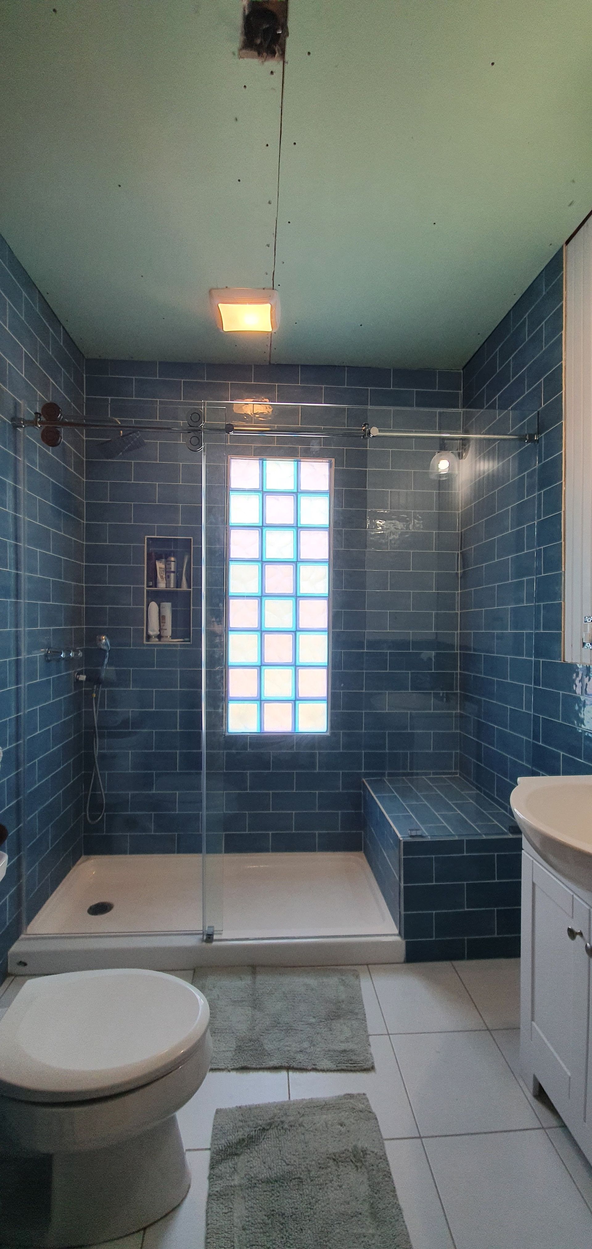 Bathroom with blue tiled shower, glass door, and white toilet. Beige tiled floor with green rug.