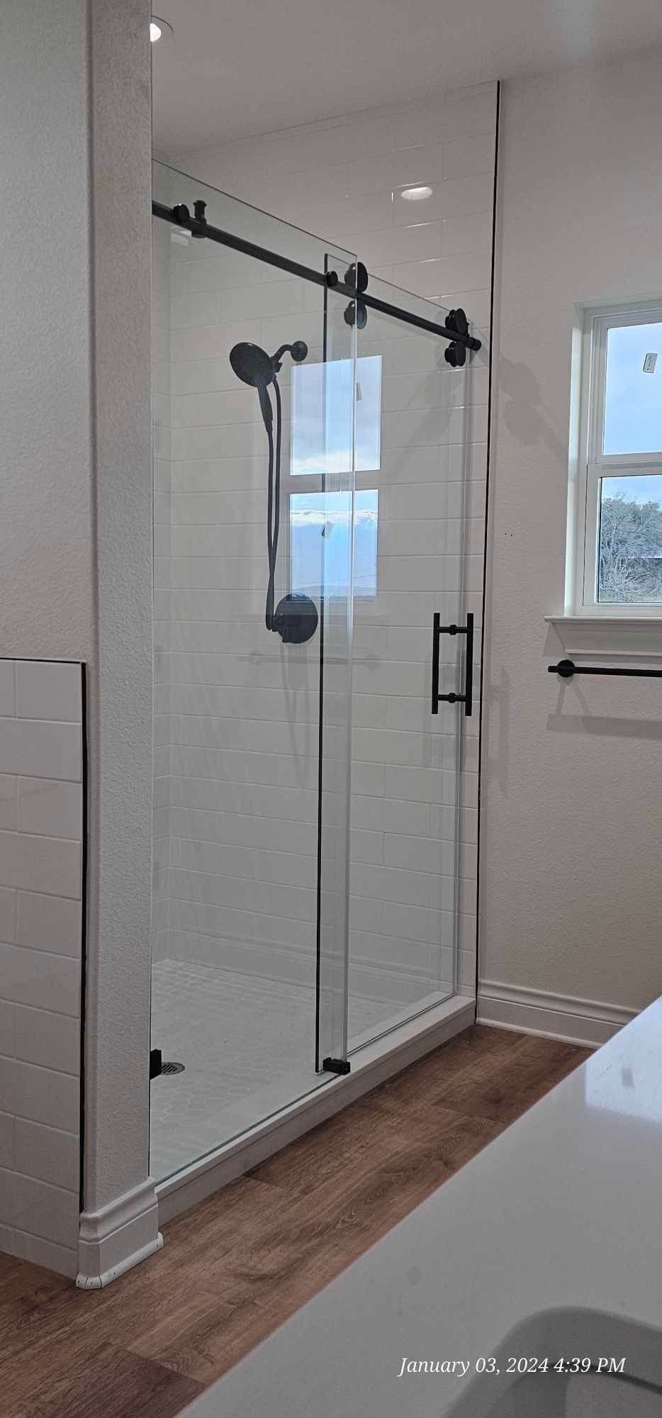 A modern bathroom with a glass shower enclosure and black hardware. The flooring is wood.