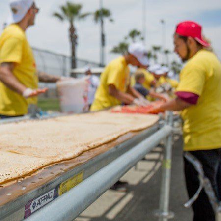 Workers in yellow shirts and pink caps handling large flat dough on a conveyor outdoors