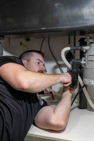 Plumber working under a kitchen sink, tightening a pipe. Black shirt, focused expression.