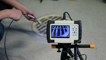 Person using a camera on a tripod to inspect a drain, monitor shows the drain's interior.