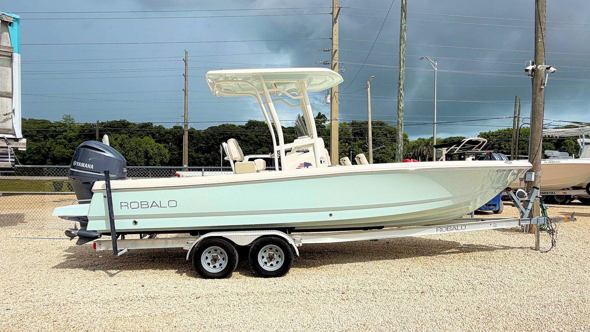 Light blue center-console boat on a trailer at a gravel lot near the water, under a cloudy sky