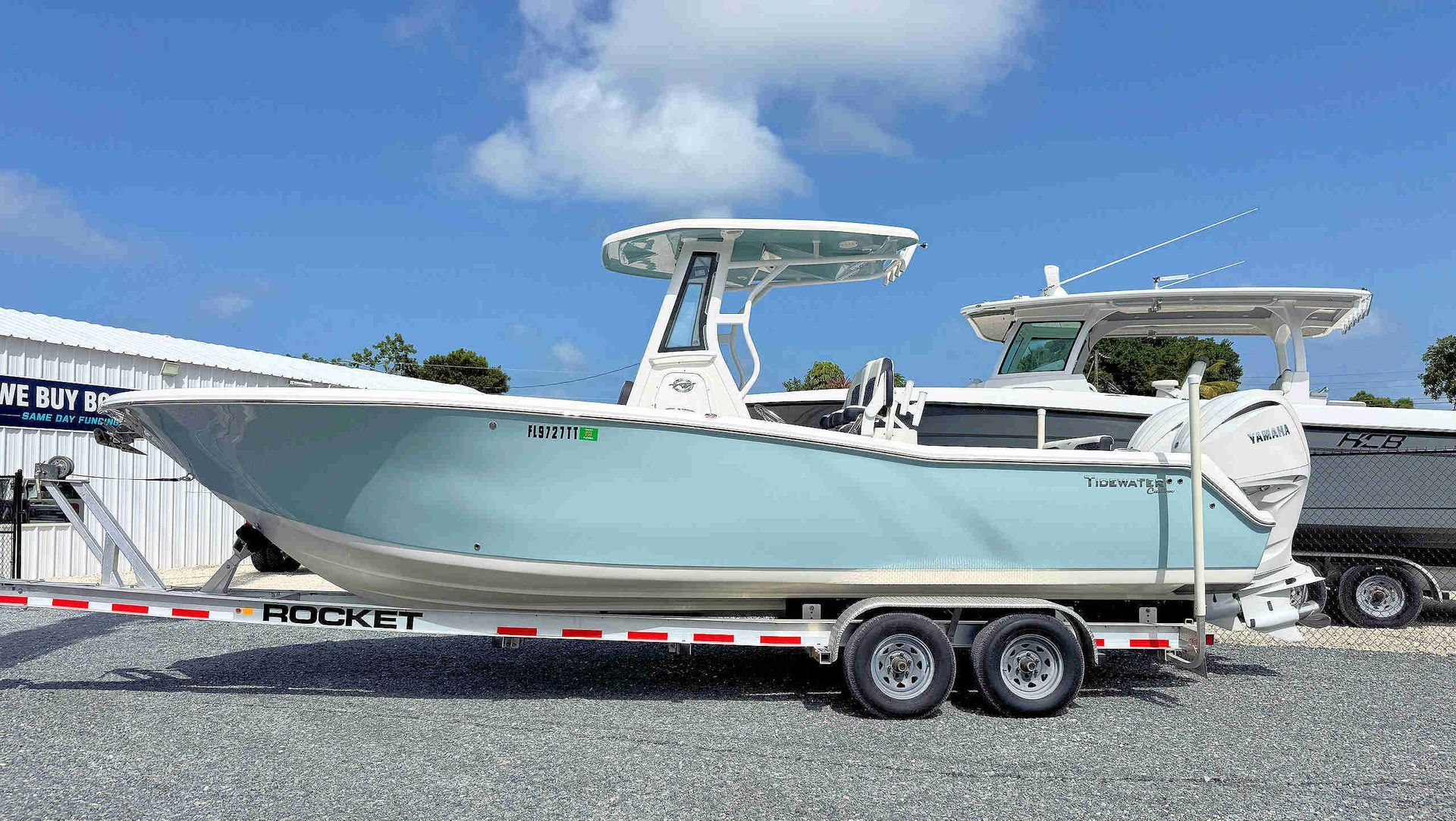 White center-console boat on a trailer in a boat yard, with blue hull stripe and twin outboard motors.