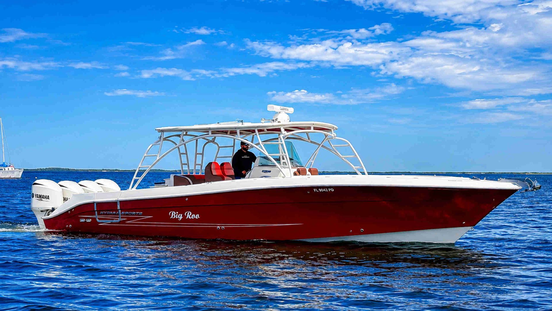 Red fishing boat on blue ocean under a bright sky, with a person aboard.