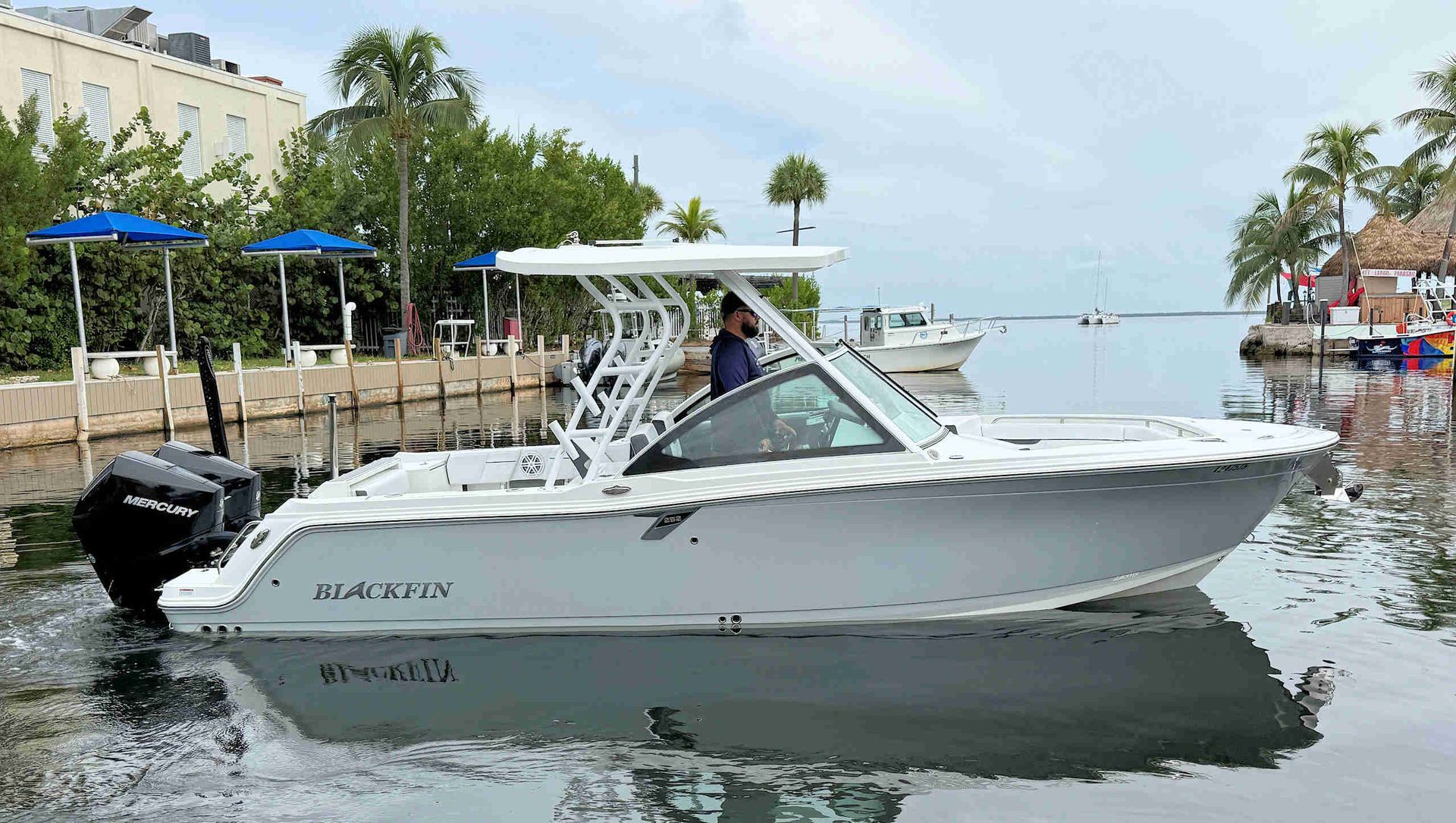 White motorboat docked on calm waterfront beside buildings and palm trees, with a person aboard