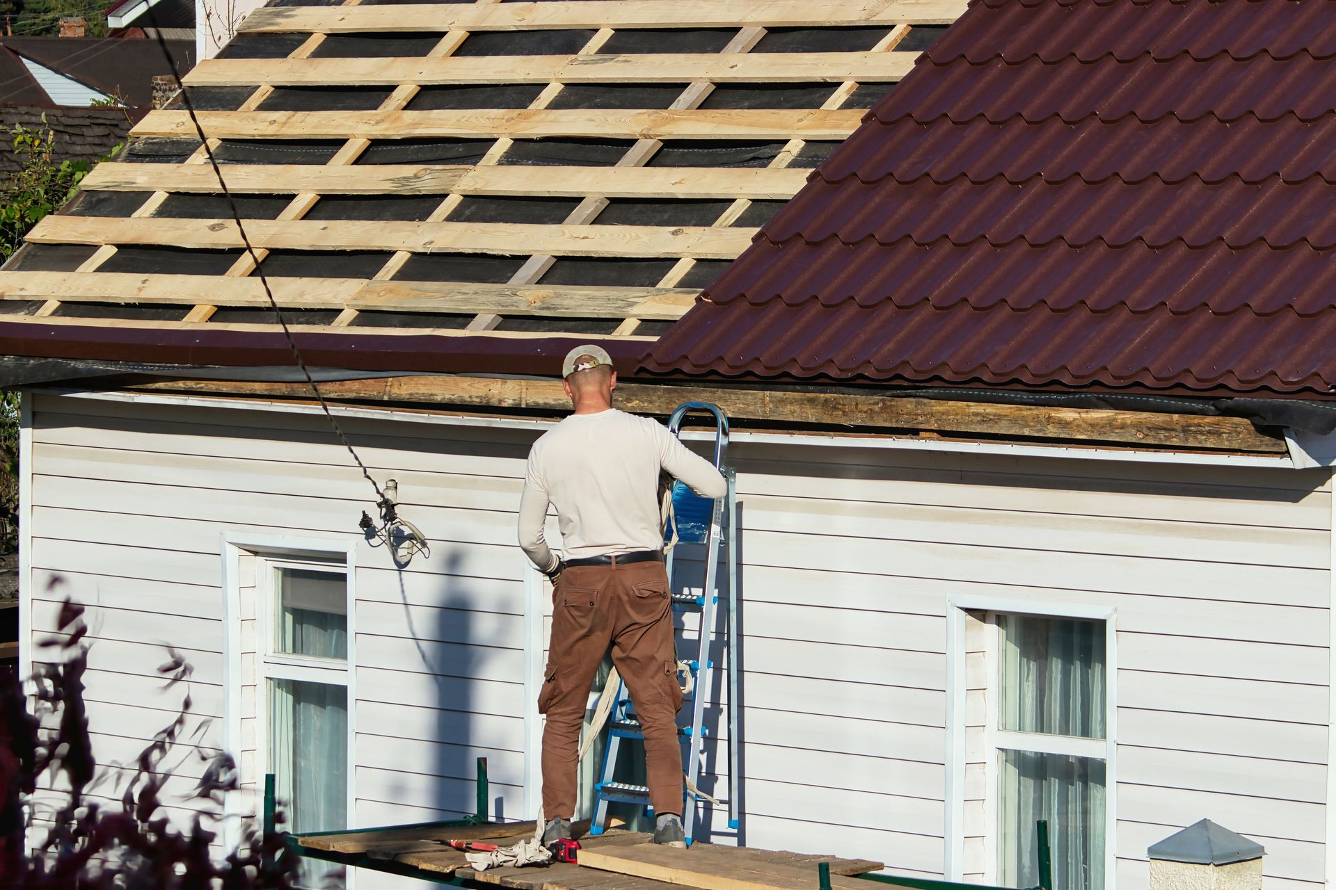 Worker standing on scaffolding repairing a roof beside a white house with exposed roof decking.