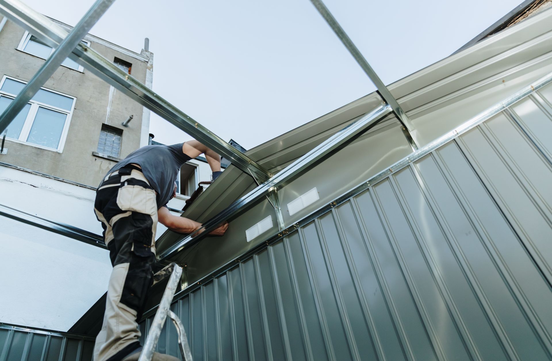 Worker on a ladder repairing a building’s roof and siding under a metal framework