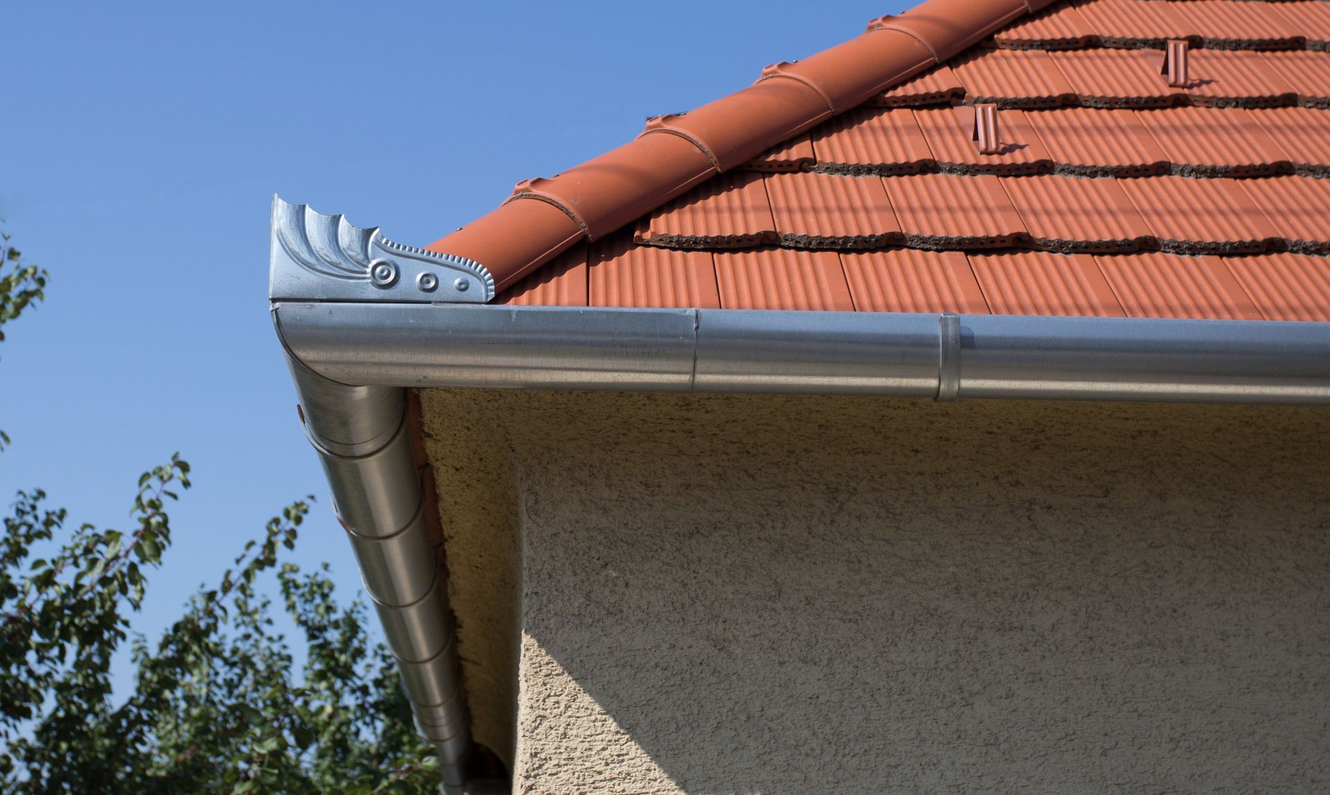 Roof corner with red tiles, gray gutter, and beige stucco wall against a blue sky.