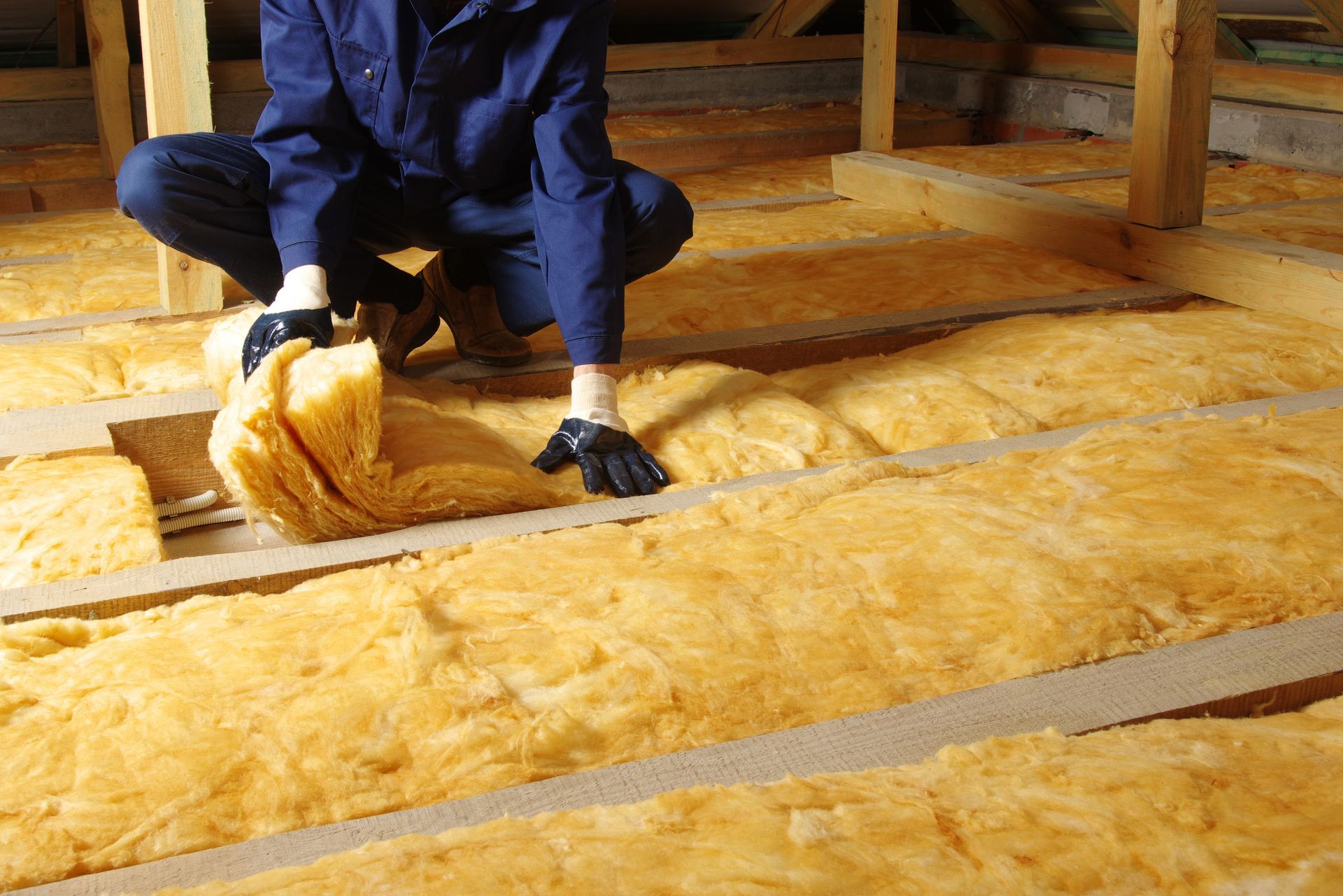 Person in blue work clothes installing yellow insulation in a wooden attic