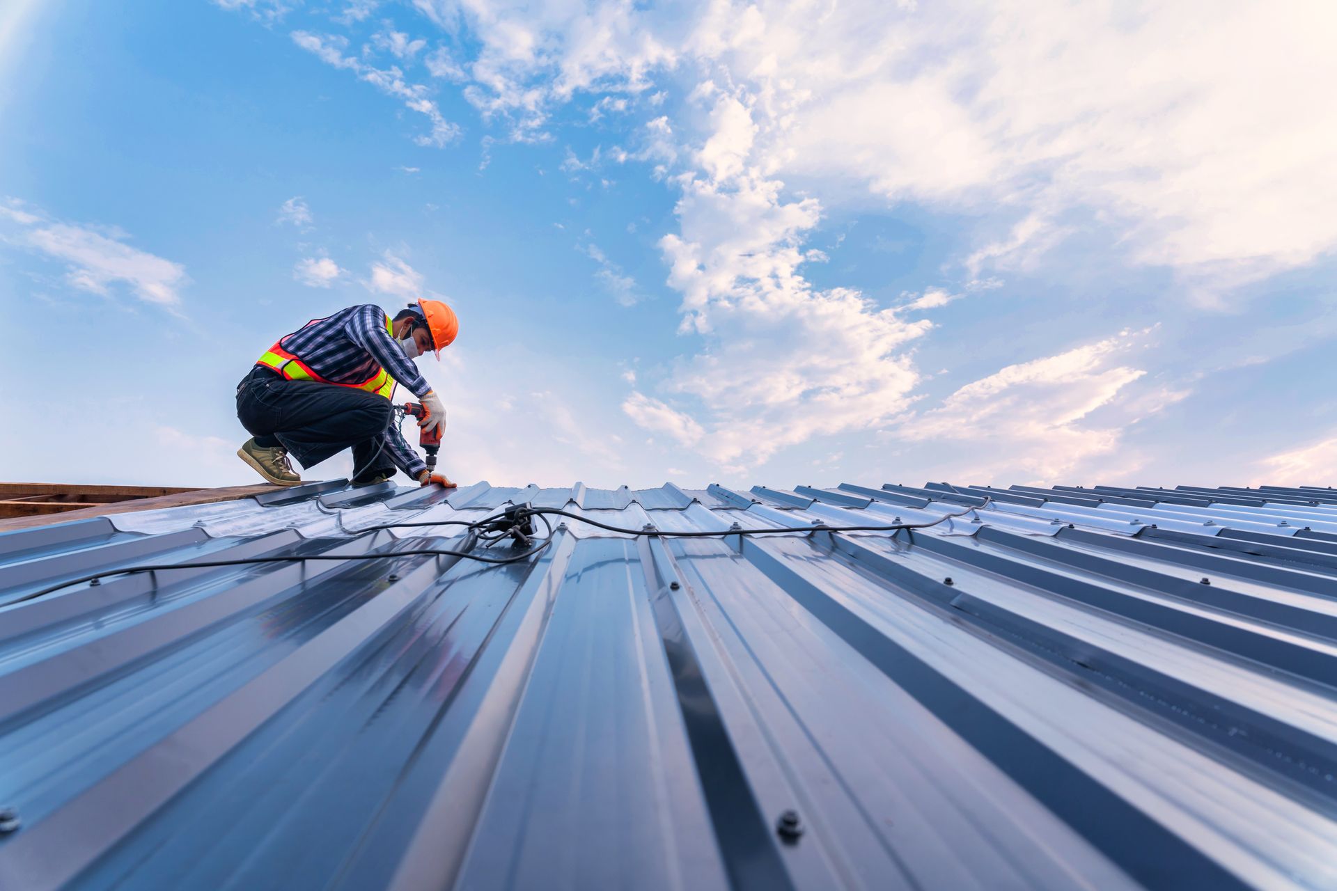 Worker in orange helmet kneeling on a blue metal roof under a cloudy sky