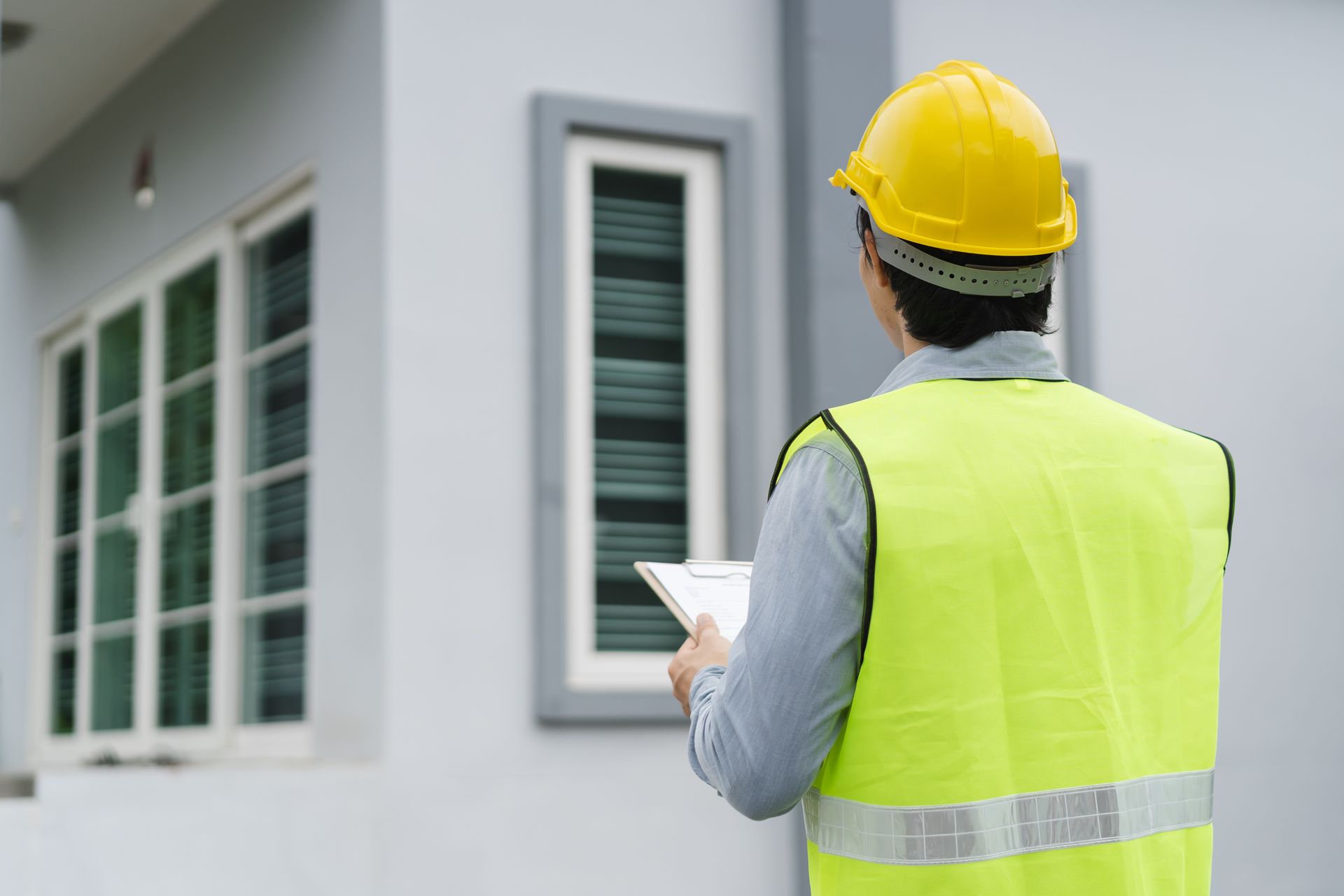 Construction worker in yellow vest and hard hat inspecting a house exterior with a clipboard