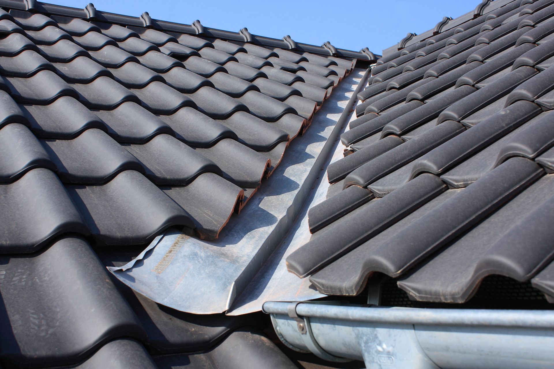 Damaged dark gray tiled roof with a bent metal valley flashing between two roof sections