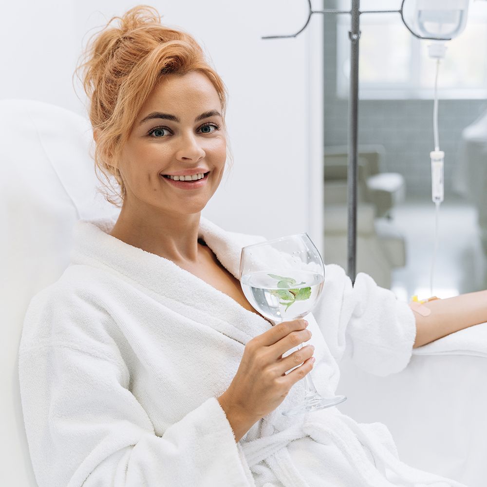 Woman in white robe, sitting, holding a glass. IV drip in background, smiling.