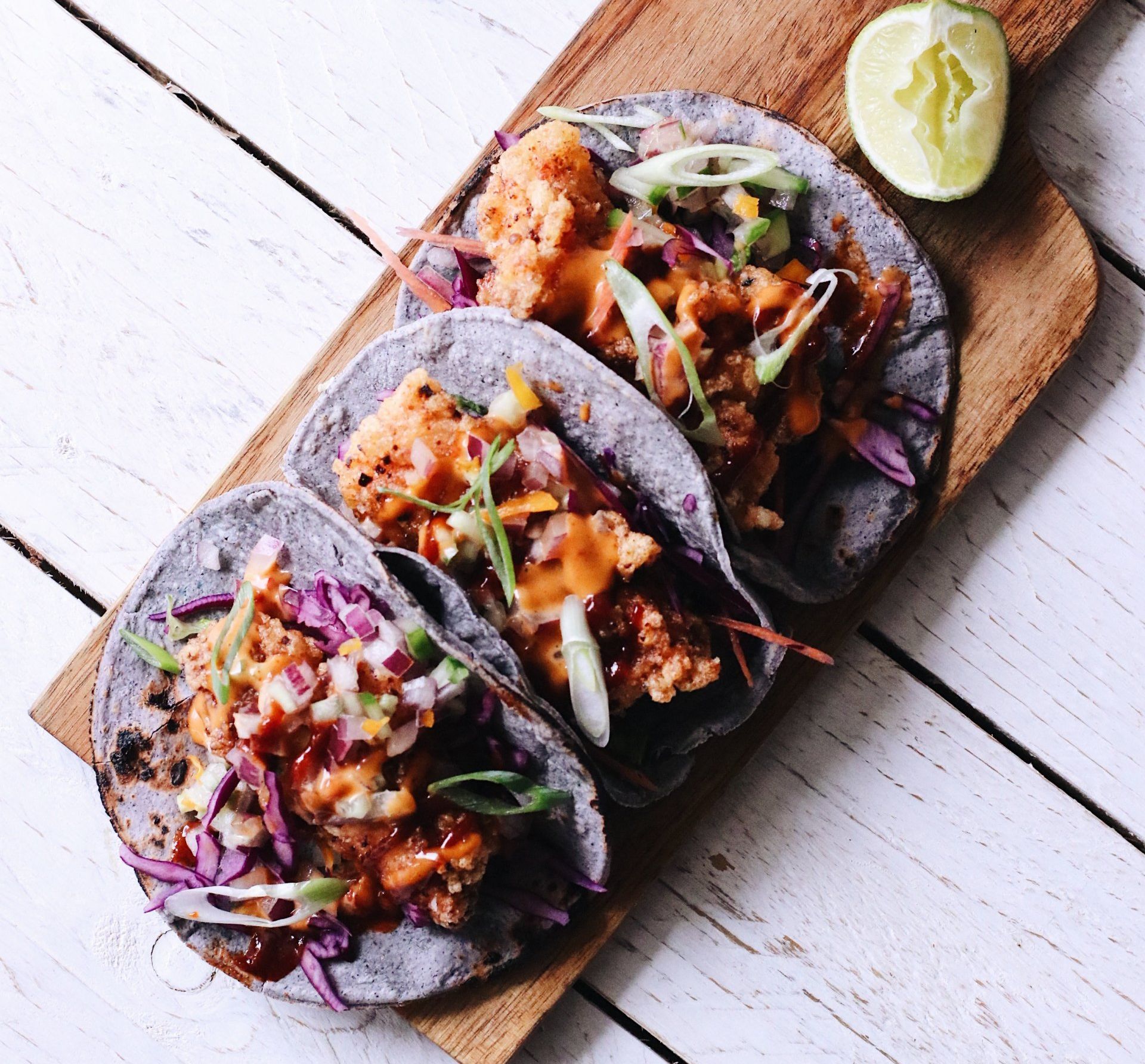 Three tacos with a purple tortilla and filled with fried fish, green onion, cabbage and hot sauce.