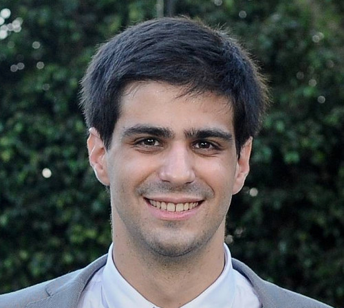 A photo of Gil in a suit and tie smiling for the camera.