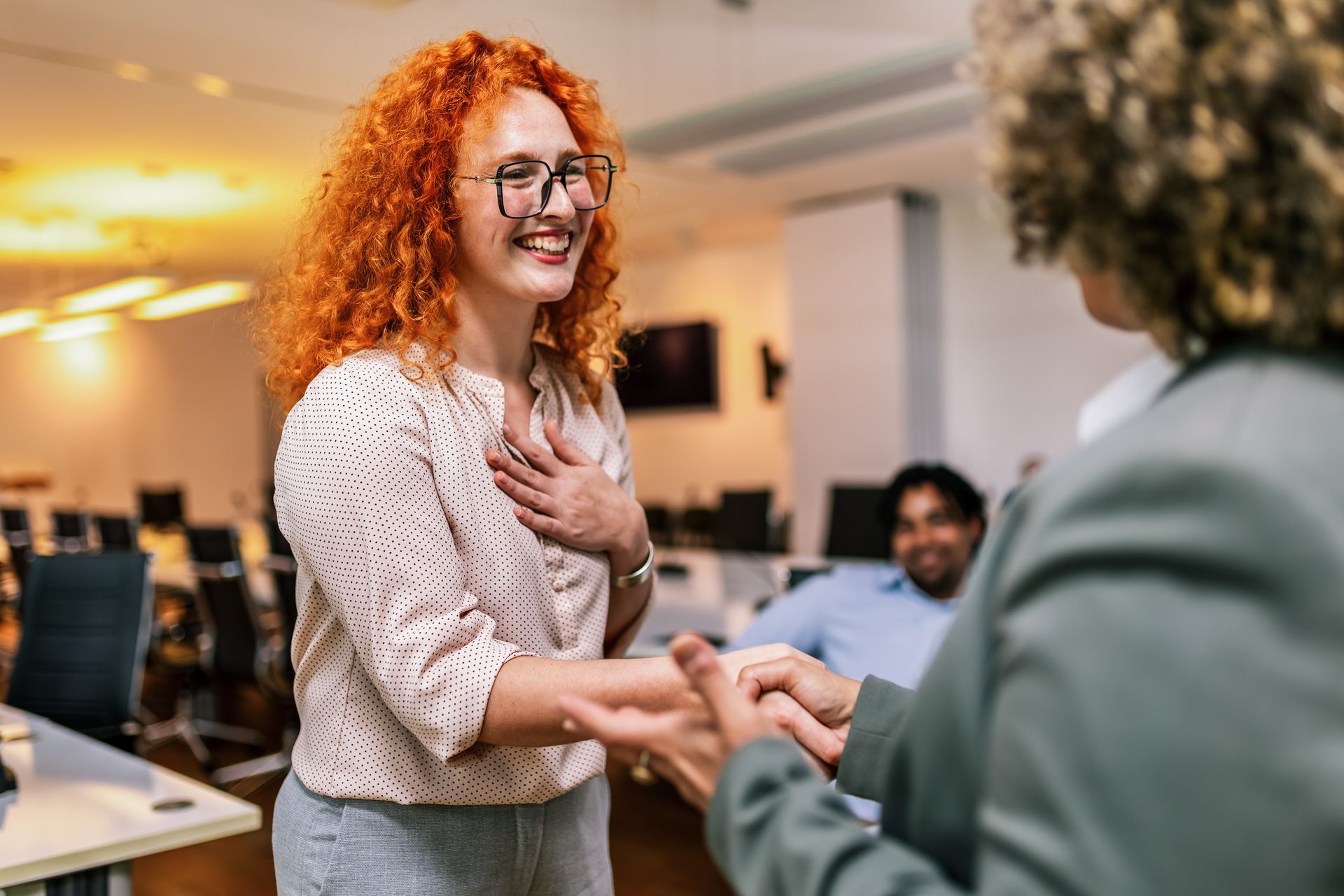Woman with red curly hair smiles and shakes hands with another woman in an office setting.
