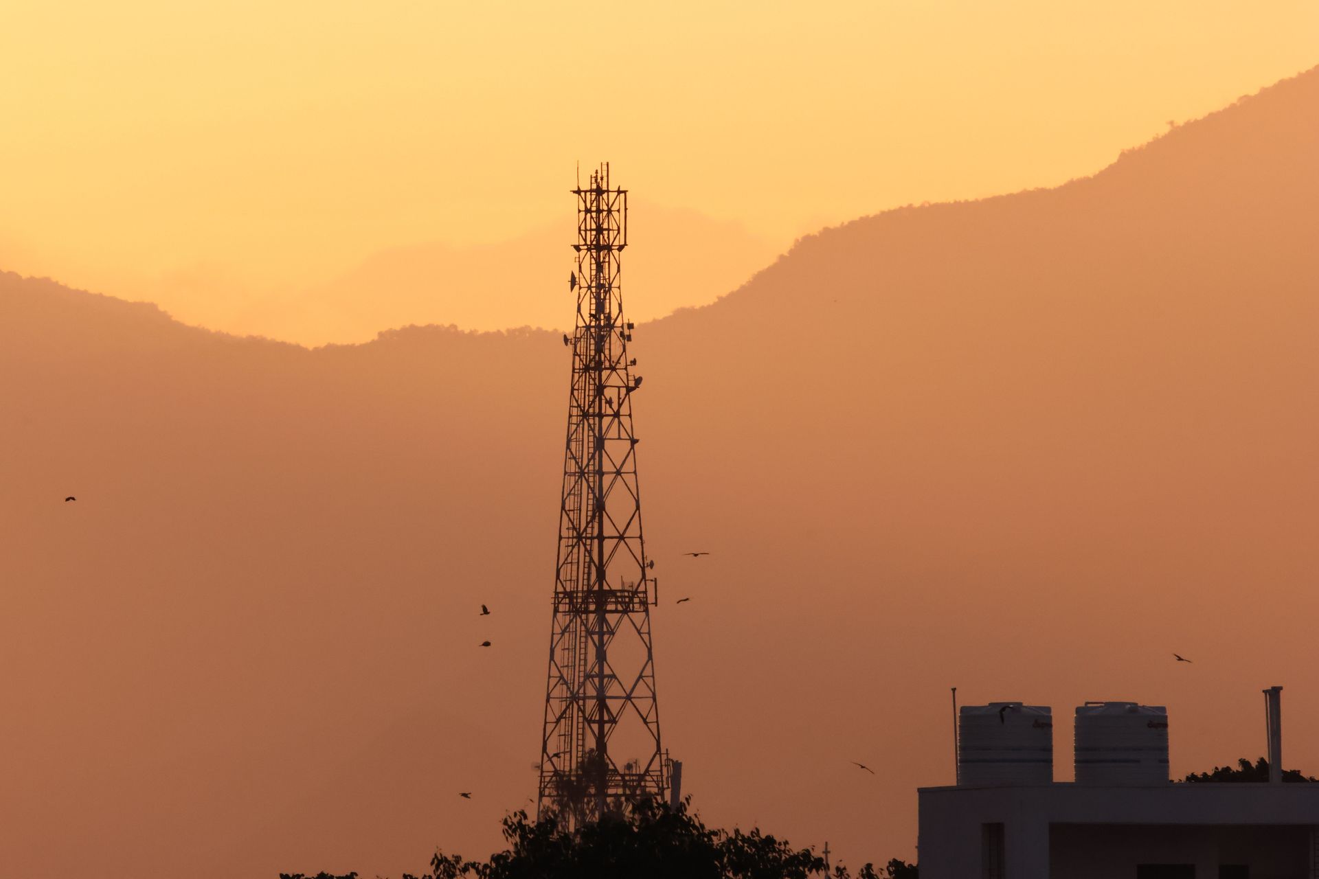 Cell tower silhouetted at sunset against hazy orange mountains and sky