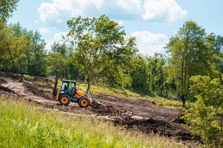 A yellow backhoe works on a muddy slope at the edge of a forest under a partly cloudy blue sky.