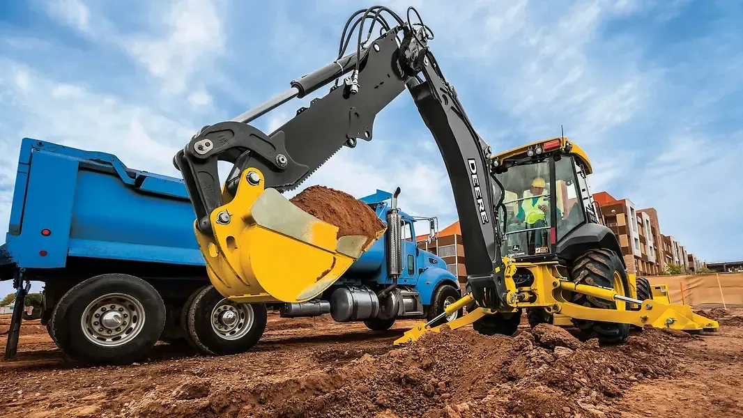 Blue dump truck and yellow excavator working in a muddy construction site, with houses in the background