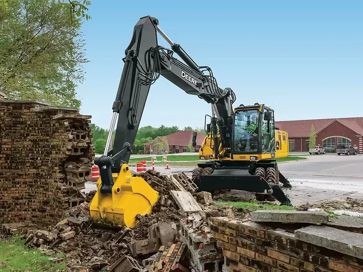 Excavator with yellow bucket clearing rubble beside a brick wall in a construction site.