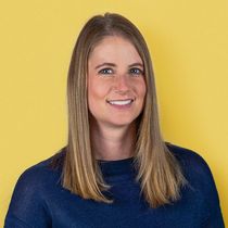 Woman with shoulder-length blonde hair smiling, wearing a navy blue sweater against a yellow background.
