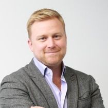 A smiling man with blond hair and a beard wearing a gray blazer over a light purple shirt.