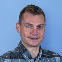 A man with short brown hair wearing a plaid shirt smiles at the camera against a light blue background.