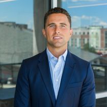 Man in a blue suit and open-collared shirt smiling, standing in front of a window with a city view.