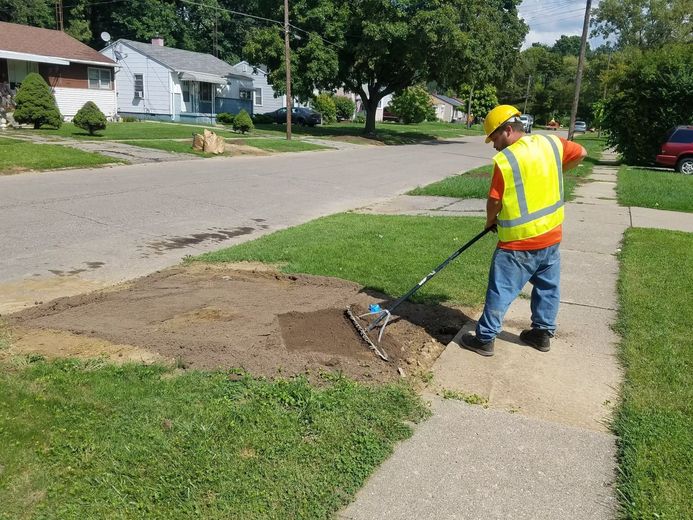Worker in a neon safety vest raking dirt beside a sidewalk in a suburban neighborhood.