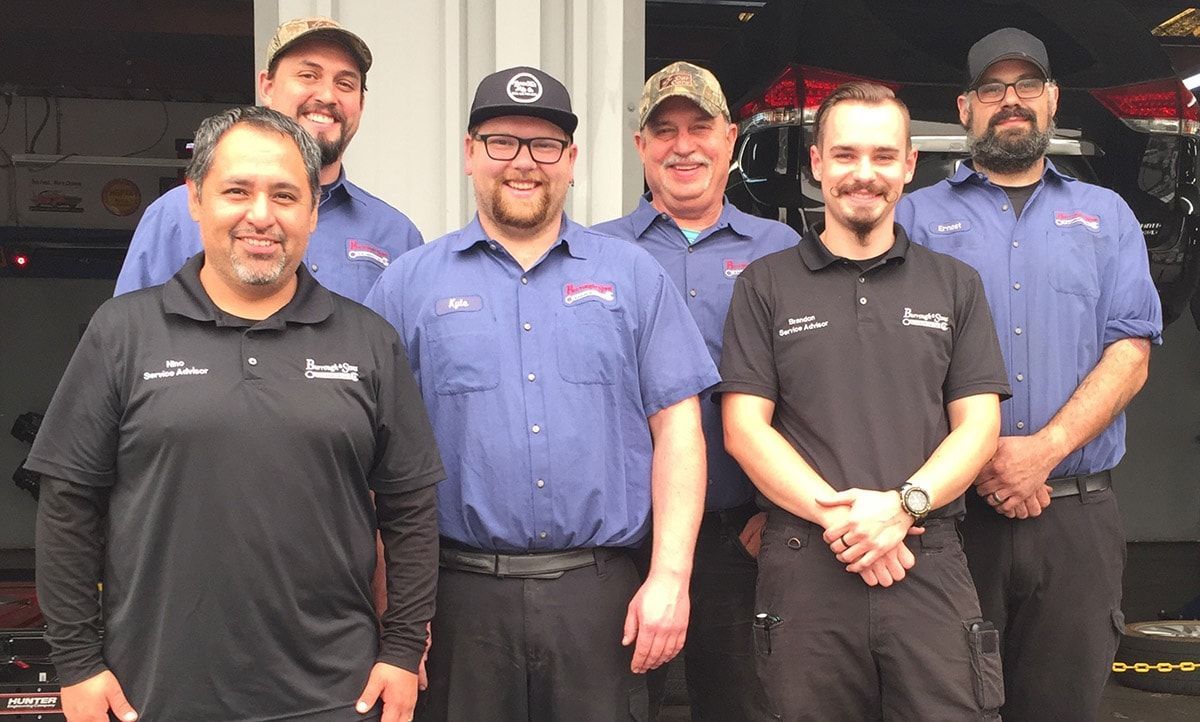 Group of six people in work shirts, smiling, posing in front of a garage. | Reliable Automotive Repair
