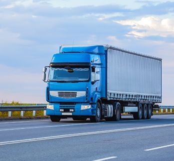 A blue semi-truck with a large white trailer driving on a highway under a cloudy sky.