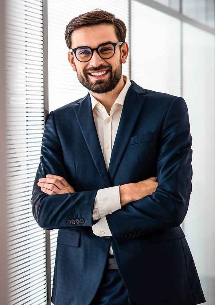 Man in blue suit, arms crossed, smiling, wearing glasses, standing near a window.