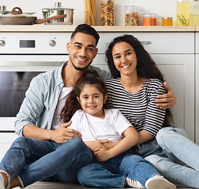 Family of three smiling in a kitchen, sitting on the floor.