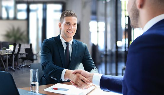 Two men in suits shake hands in an office, one smiling, at a desk.