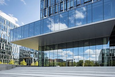 A wide-angle shot of a modern glass office building with a large, protruding cantilevered entrance over stone steps.