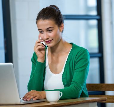 Woman on phone, using laptop, drinks coffee, green sweater, smiles.