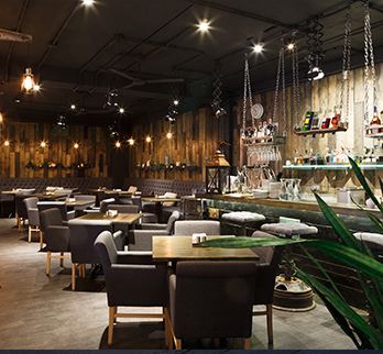 A dimly lit restaurant interior with dark walls, wooden accents, cushioned dining chairs, and a bar with hanging shelves.