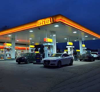 A lit Shell gas station at twilight with several parked cars and a person standing by a pump under the canopy.