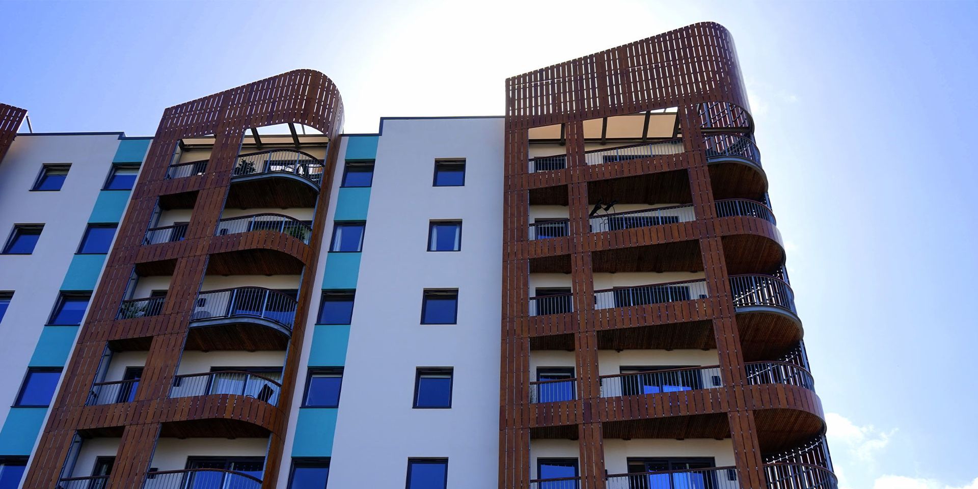 A multi-story building with white, teal, and brown facade against a bright blue sky.