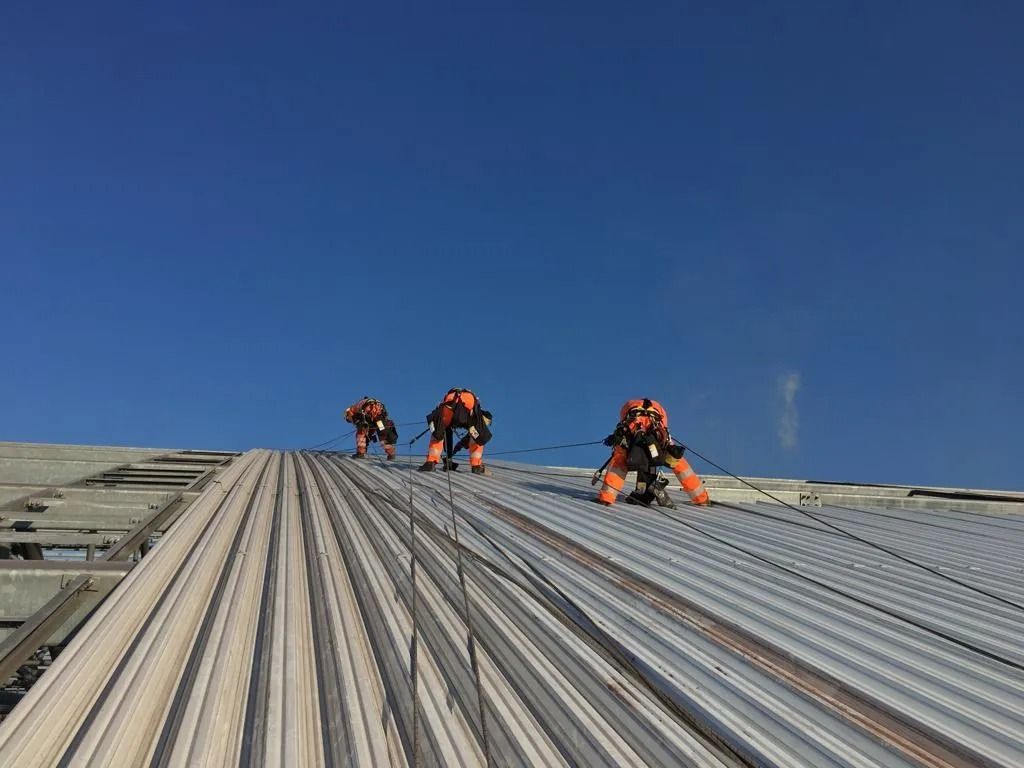 Tre operai in tute arancioni su un tetto di metallo, che fissano le corde, con il cielo azzurro sopra di loro.