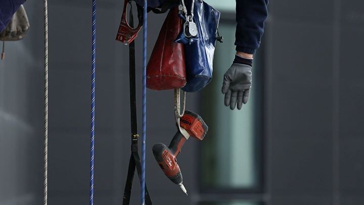Persona che lavora in quota, appesa con degli attrezzi; mano guantata grigia che allunga la mano verso una borsa portautensili rossa e blu con un trapano elettrico penzolante.