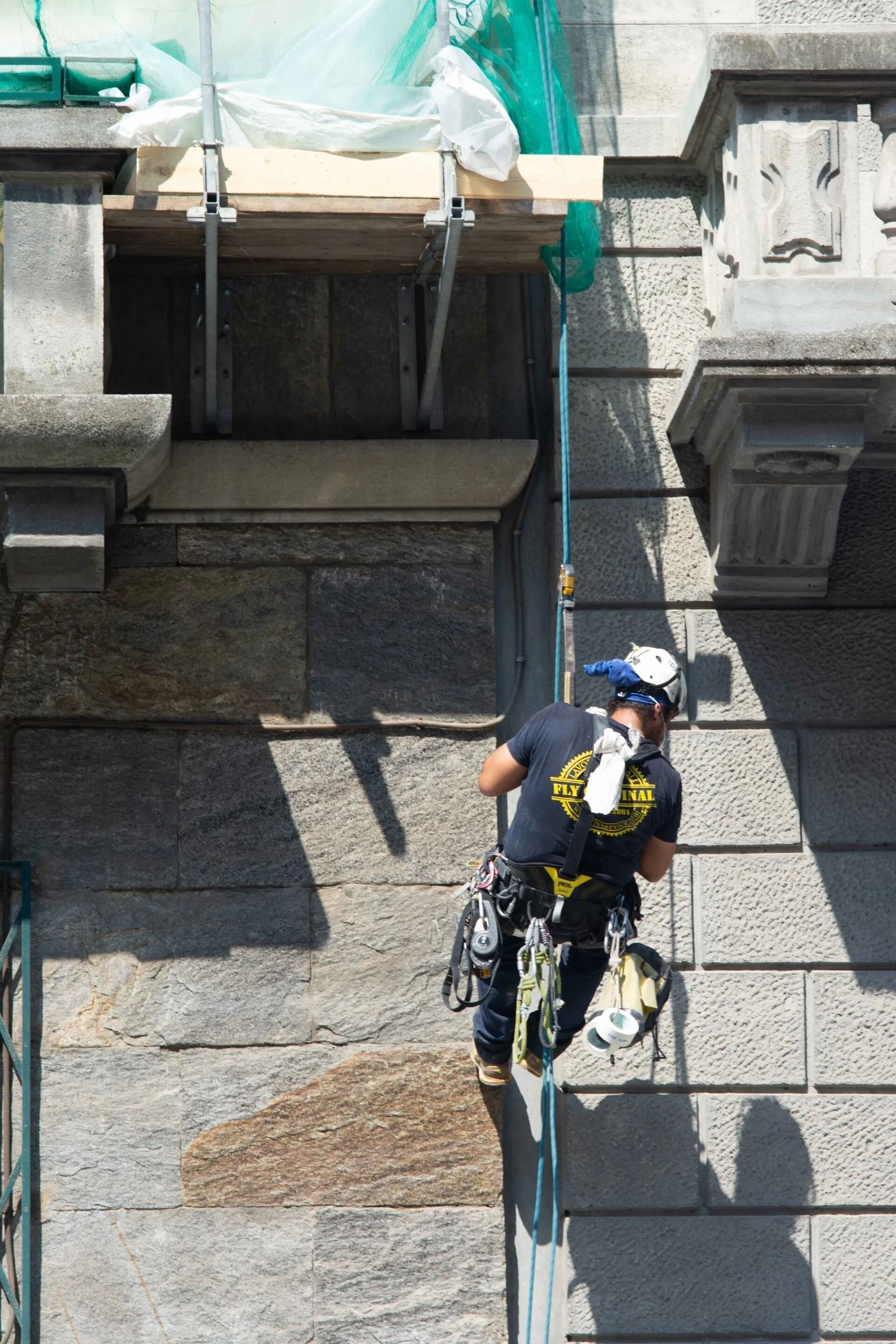 Operaio edile che si cala in corda doppia da un edificio in pietra, utilizzando dispositivi di sicurezza. Impalcature verdi e piattaforma in legno.