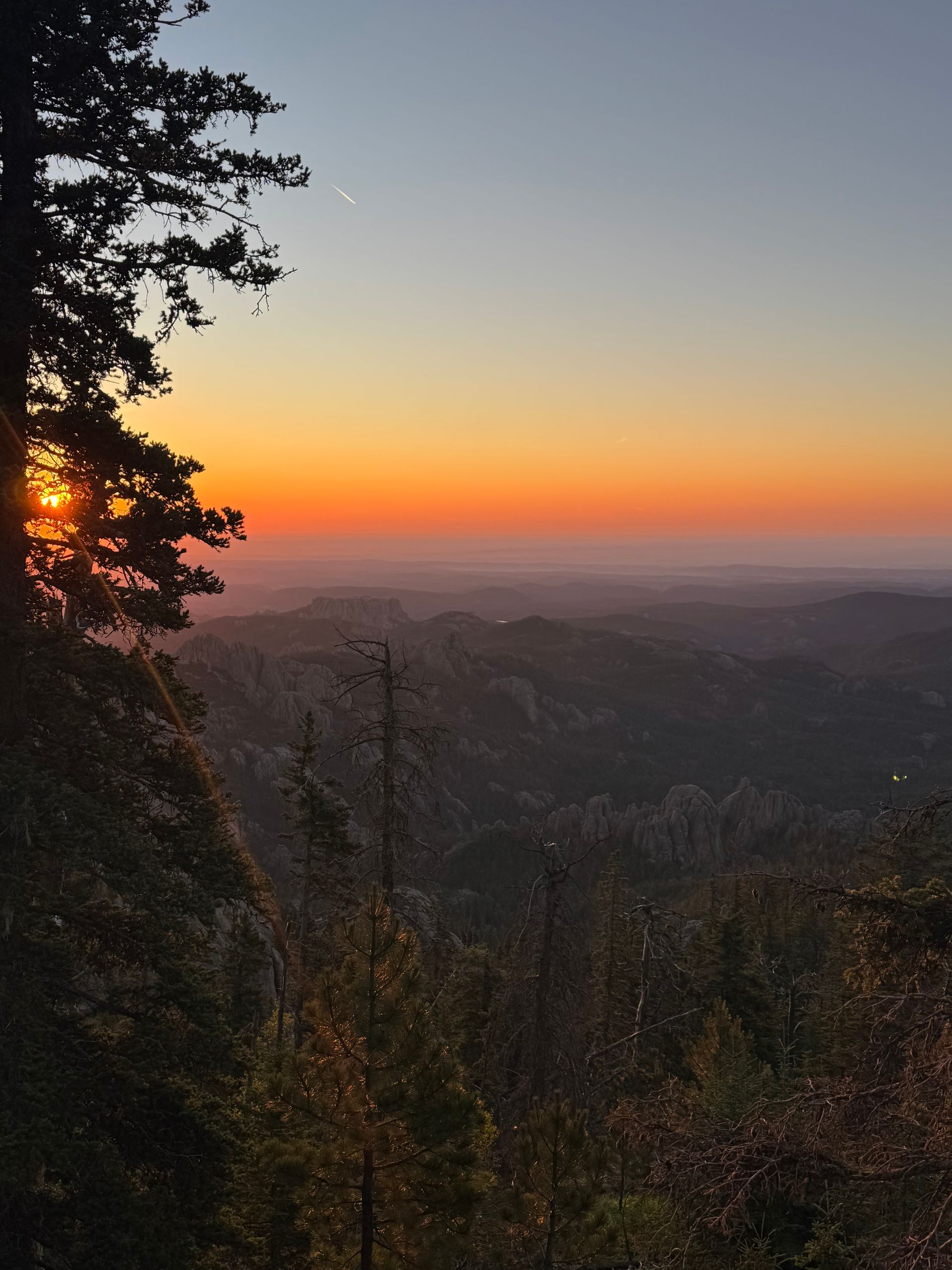Sunset over hazy hills, with silhouetted trees and a glowing orange horizon