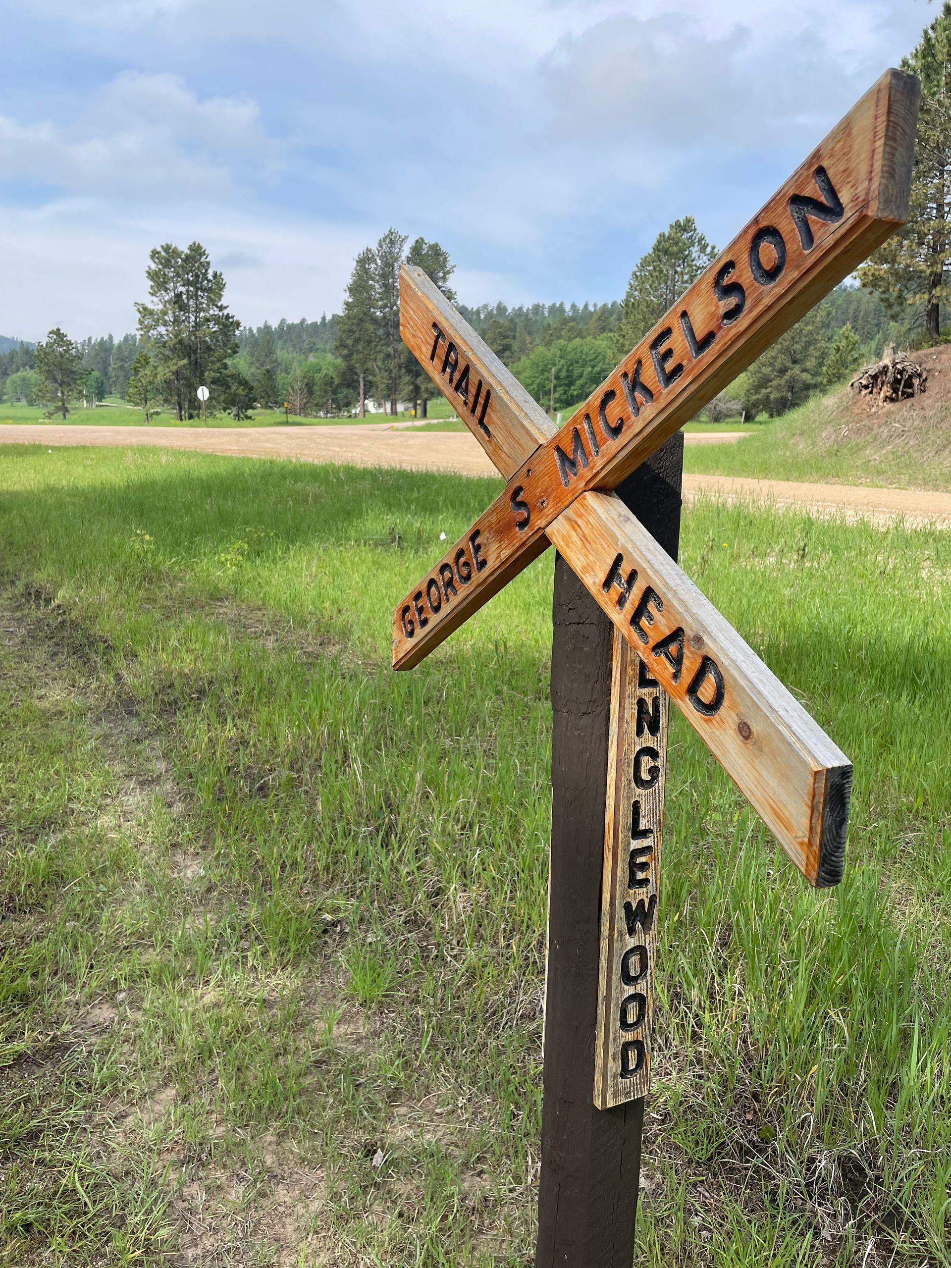 Rustic wooden trail sign at a grassy roadside intersection under a clear sky.