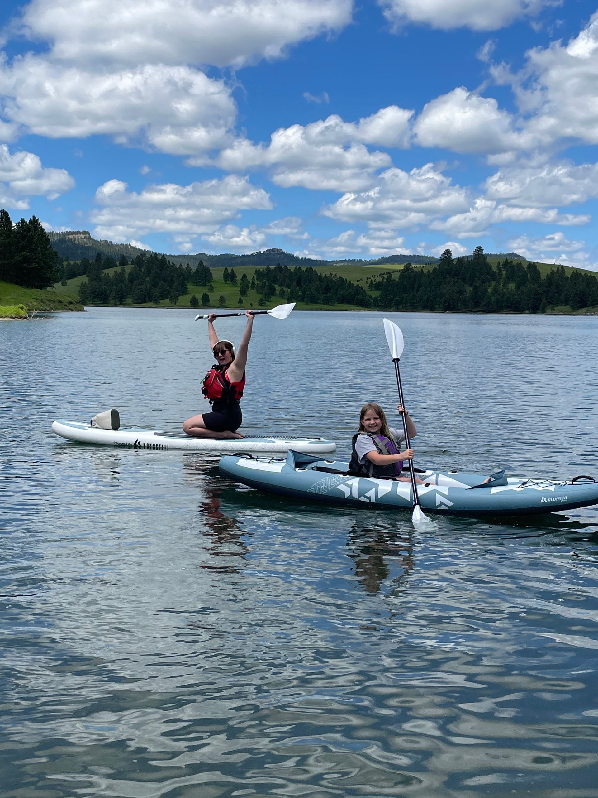 Two people on kayaks on a lake, one raising arms under a cloudy blue sky.