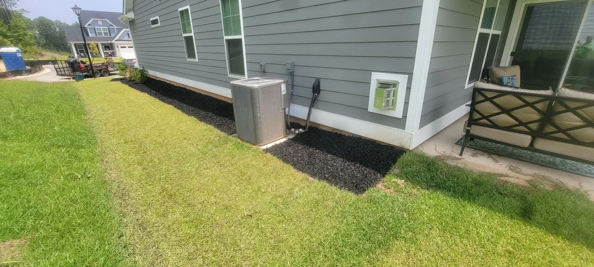Side of a gray house with air conditioner and mulch bed, with green lawn.